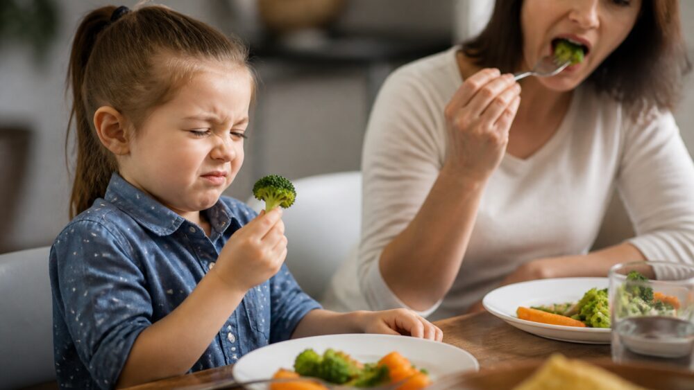 کودکی پشت میز غذا با حالت تردید به بشقاب بروکلی و سبزی های برگ سبز نگاه می کند