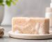 یک قالب صابون کرم‌رنگ با دانه‌های ریز لایه‌بردار روی حوله سفید، کنار کاسه‌ای از نمک دریایی و برگ سبز
