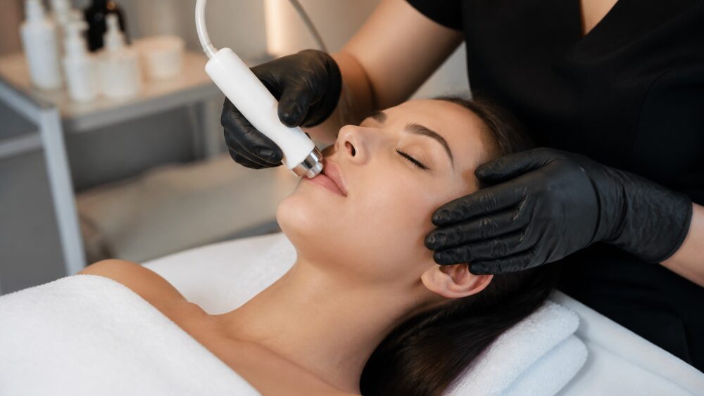 متخصص پوست در حال ماساژ آرام صورت مراجعه کننده روی تخت درمانی، با محصولات مراقبتی در پس زمینه