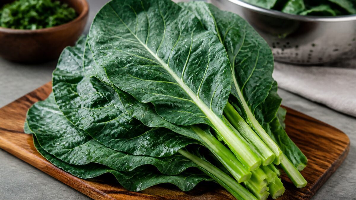 دسته‌ای کلم کولارد تازه با برگ‌های سبز تیره روی تخته چوبی در آشپزخانه روشن