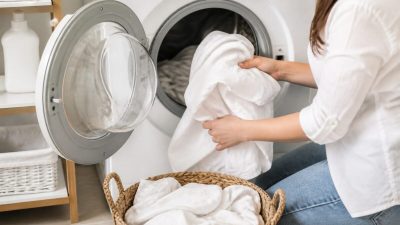 دست‌ها در حال قرار دادن لباس‌های سفید تمیز داخل ماشین لباسشویی در فضای رختشویخانه روشن