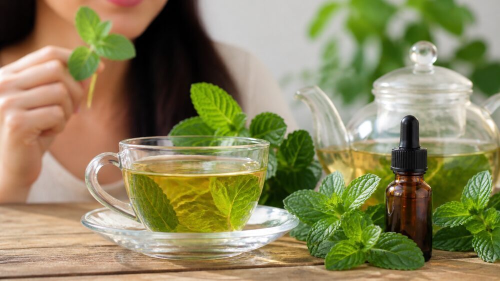 فنجان دمنوش نعناع کنار برگ های تازه نعناع و شیشه کوچک روغن گیاهی روی میز چوبی