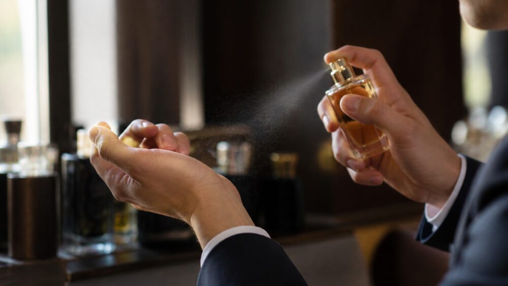 دست در حال اسپری کردن عطر از شیشه کوچک روی مچ دست، کنار چند شیشه عطر با اندازه های متفاوت