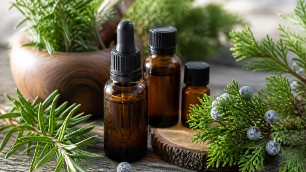 بطری شیشه ای روغن رزماری کنار شاخه های تازه رزماری و حوله سفید روی میز چوبی