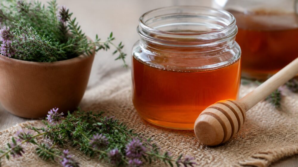 شیشه دربسته شربت آویشن با عسل کنار قاشق چای خوری و شاخه های آویشن روی میز آشپزخانه