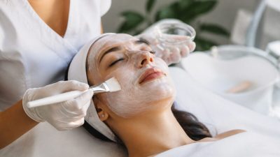 متخصص پوست با برس نرم ماسک مراقبتی را روی صورت زنی در فضای روشن و تمیز می‌زند