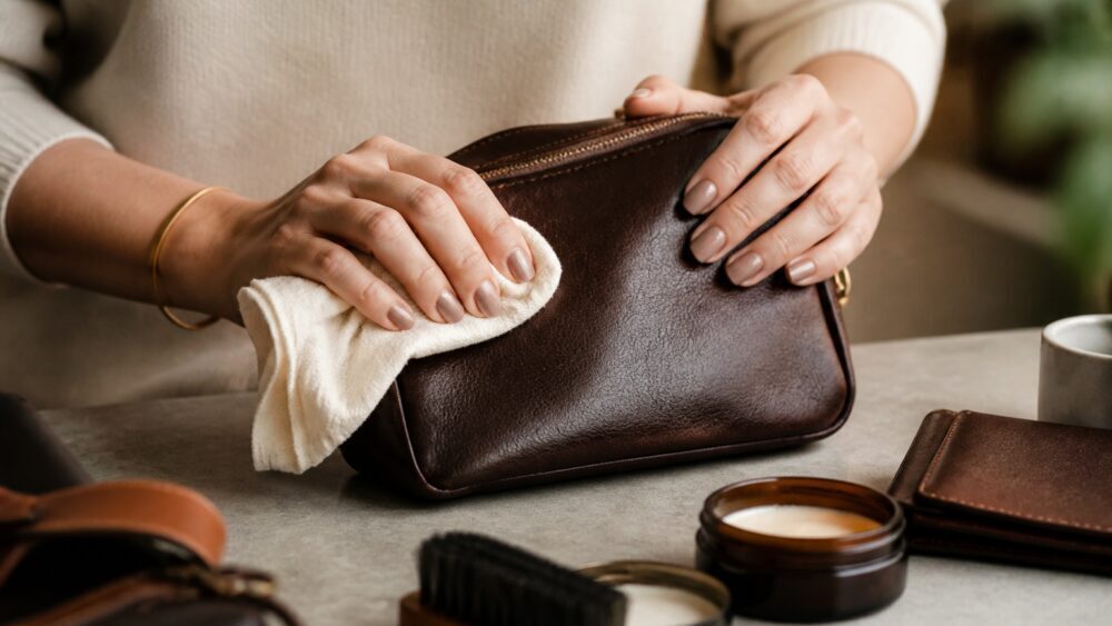 دستی در حال پاک کردن یک کیف چرمی قهوه ای با دستمال نرم روی میز، دور از نور مستقیم